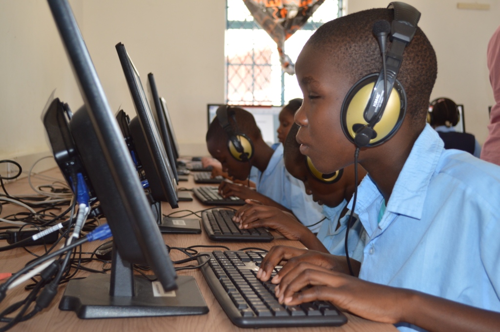 A picture of a visually impaired learner wearing headphones and seated in front of a desktop computer while typing with his fingers on the keyboard. He's wearing a light blue shirt and inside a computer lab for the blind.