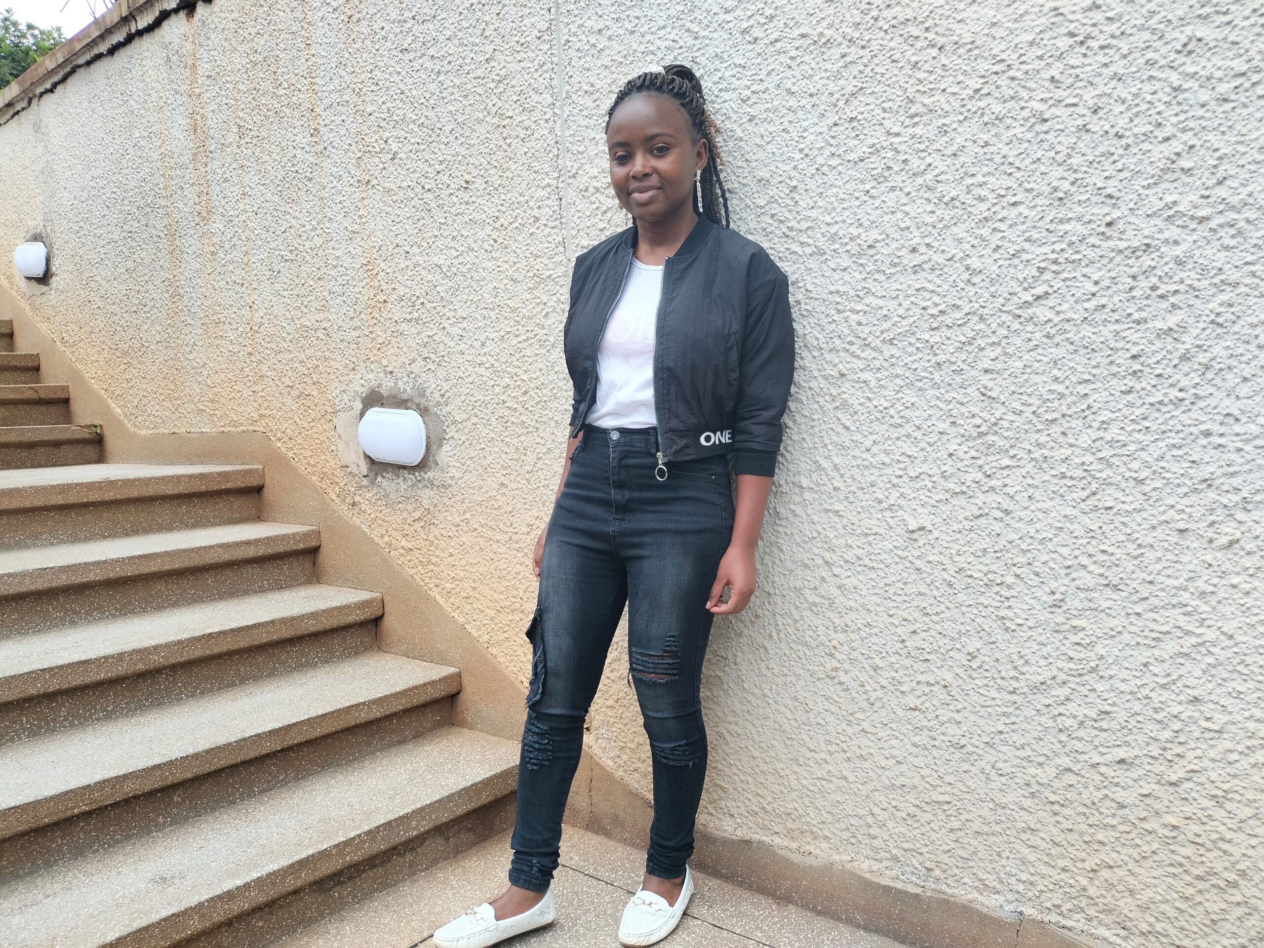 A picture of Brenda Mwende smiling and leaning against a wall. She is wearing a white shirt, a black jacket, blue jeans and white shoes.