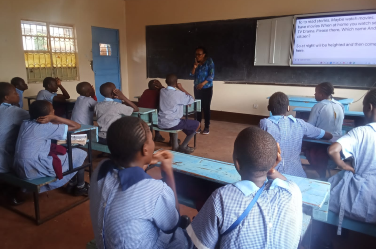 A teacher at Kambui School for the Deaf is interacting with students in a classroom setting. A screen displays text related to the lesson.
