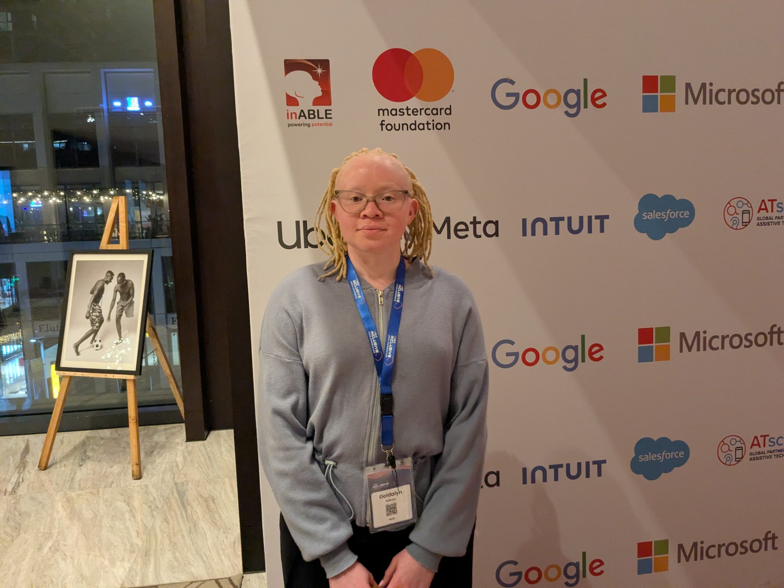 A photo of Goldalyn with light-colored dreadlocks stands in front of a white backdrop filled with the logos of corporate sponsors like Google, Microsoft, and Meta. She is wearing glasses and a light gray zippered sweater. A conference lanyard with a nametag is around her neck. To her left, an easel holds a framed black and white photograph of two people playing soccer. The background is a blurry conference hall with lights and windows.