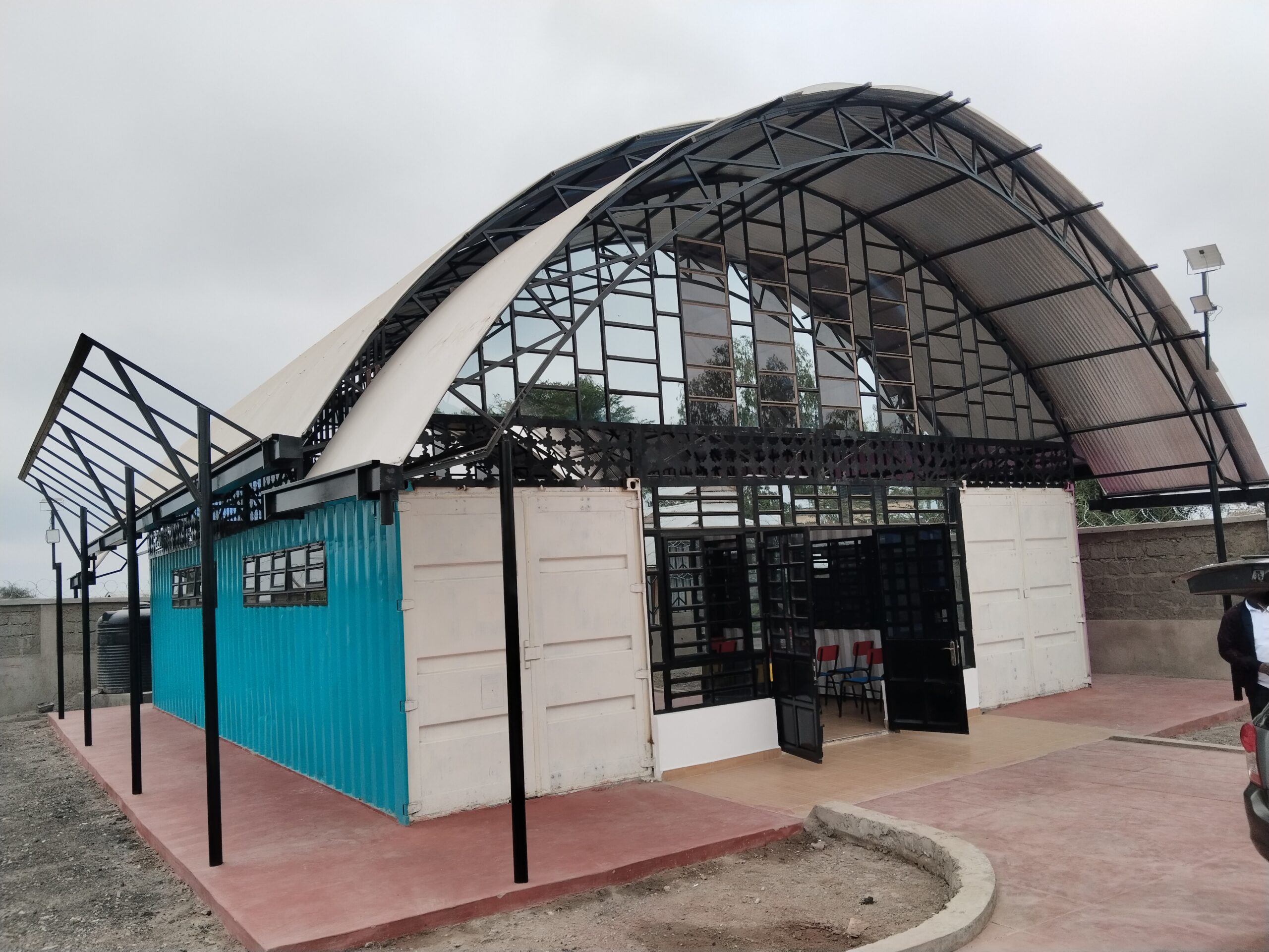 A picture of Isinya Digital Hub, a modern building with a curved metal roof, glass windows, and white and blue walls. The design is industrial and innovative against a cloudy sky.