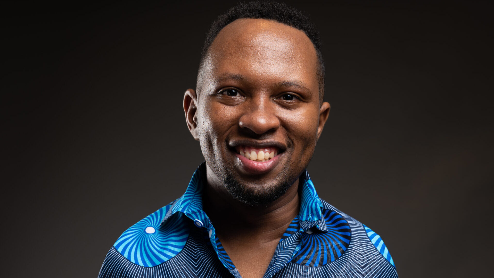 A picture of Sammy wearing a blue patterned shirt with geometric circles stands against a dark background.