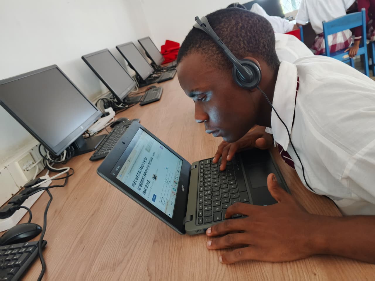 A student wearing headphones leans very close to a laptop screen while working in a computer lab. Several desktop computers are arranged on the desk beside him, and other students in school uniforms are seated in the background.