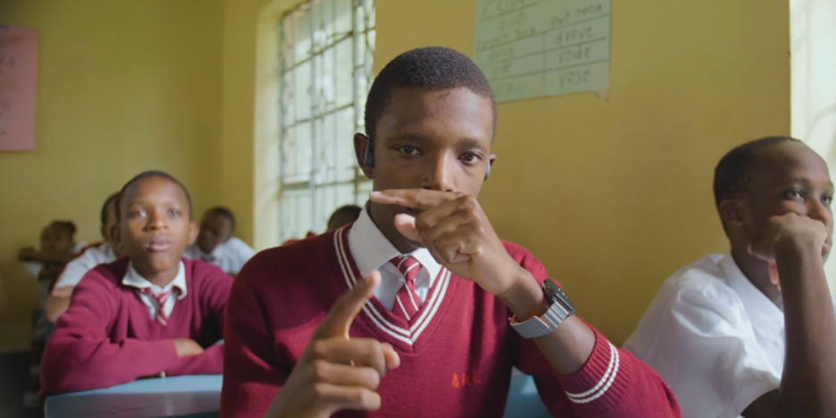 Close-up of a student in a maroon sweater, tie, and smartwatch, looking forward and touching his mouth in a pensive gesture.