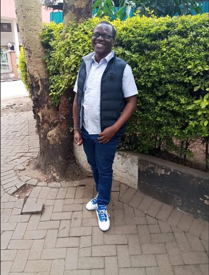 Maurice is smiling and stands outdoors beside a tree and a neatly trimmed green hedge. He is wearing glasses, a short-sleeved light blue shirt, a black sleeveless vest, dark blue trousers, and white sneakers with blue accents. The setting appears to be a paved walkway in a residential or garden area.